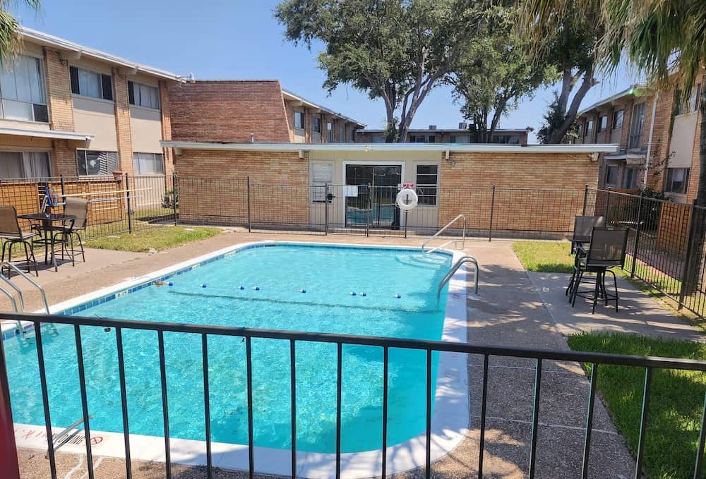 A gated swimming pool with two tables and four chairs, grass, and trees, surrounded by apartment buildings.